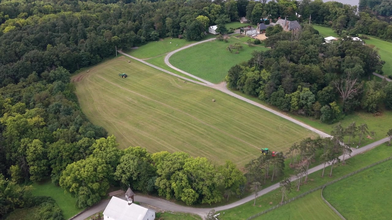 An aerial drone shot capturing agricultural tractors driving across open farm fields surrounded by green pastures and countryside trees.
