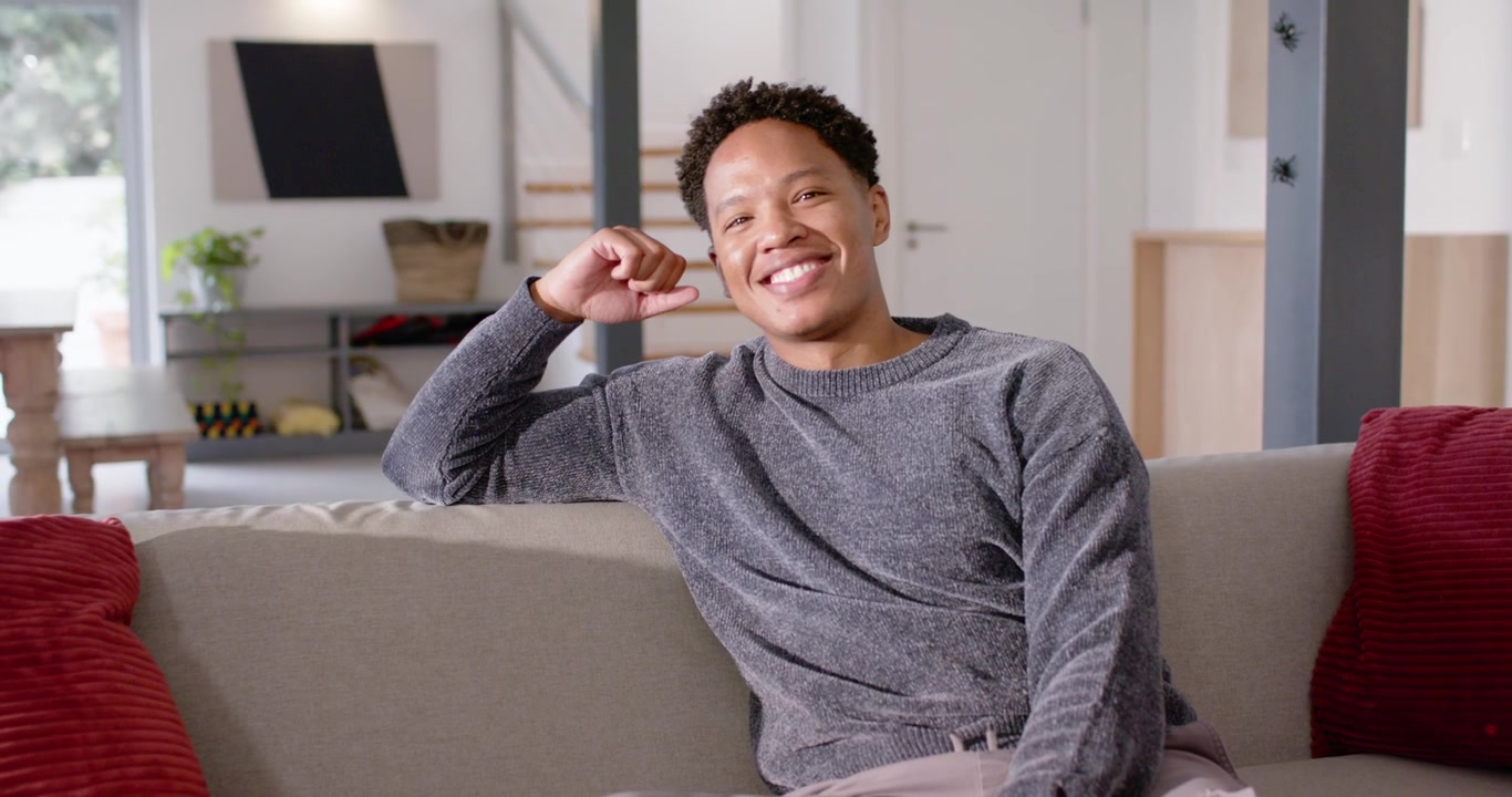African American man sitting on sofa at home after camera zoom, resting head on hand, smiling
