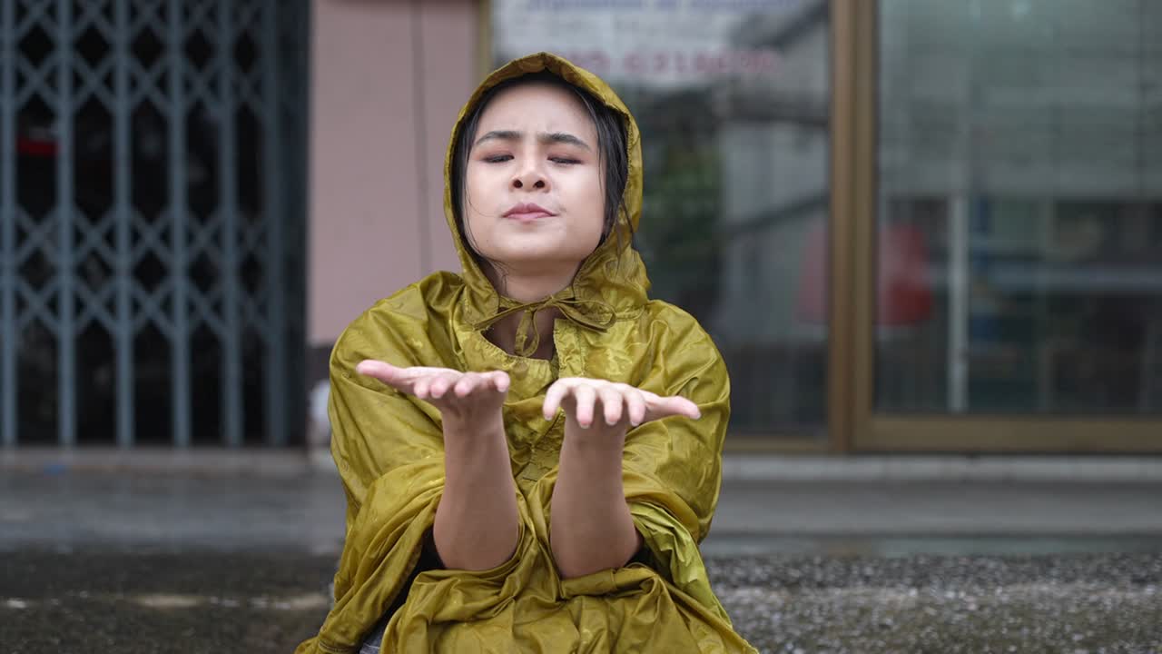 A young female freelance teacher boring squatting under roof waiting for rain to stop, being late for work, uncomfortable of getting wet in rainy day, reach out hands touching raindrops on palms,