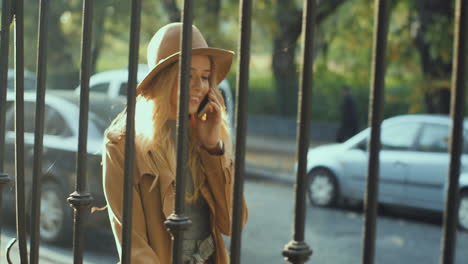 cheerful smiled blonde young woman weating a hat and coat walking behind a fence and talking on the phone in the street