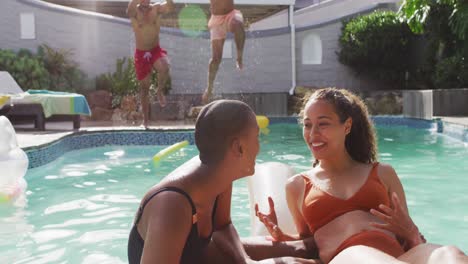 Two-diverse-female-friends-talking-in-swimming-pool,-their-male-friends-jumping-into-water