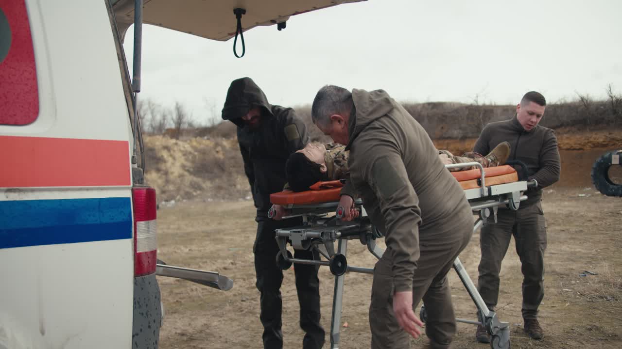 tres hombres confiados y militares en uniformes verdes pusieron a un militar herido en uniforme de camuflaje en una trampa con una muñeca en una ambulancia durante un ejercicio de emergencia médica en un campo de entrenamiento militar