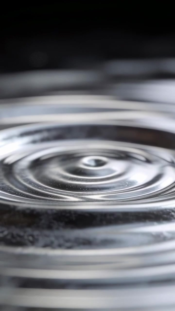 Video verticale: Goccia che impatta sulla superficie dell'acqua, formando increspature in una pozza poco profonda, con bagliori.