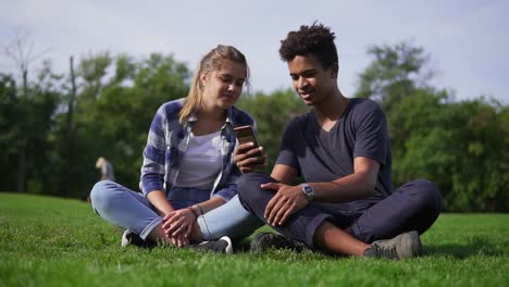 mixed race couple sitting on the grass and selects pictures in their smartphone. young people smile and laugh. cute friends