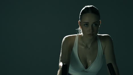 medium closeup of a sweaty woman training on a bike, seen from waist up as the camera pans left to right, revealing her focused eyes, glistening chest, and sculpted upper body in dramatic lighting