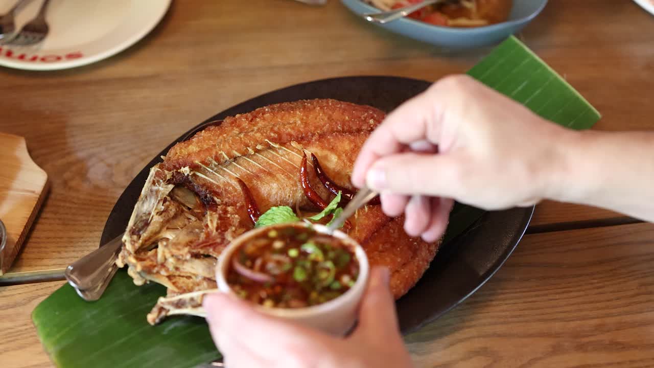 Hand pours spicy herb sauce onto fried fish on banana leaf in bright restaurant setting