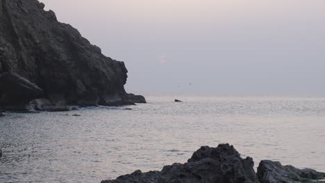 Ruhiger,-Wolkiger-Sonnenaufgang-In-Der-Nähe-Eines-Felsigen-Strandes,-Während-Möwen-über-Wasser-Fliegen,-Während-Plötzlich-Ein-Mensch-Auf-Einen-Felsen-Springt