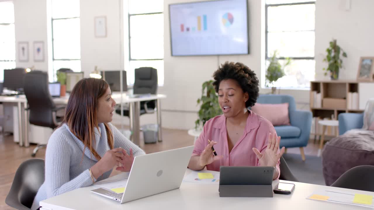Diverse business female colleagues in discussion using tablet and laptop in office, slow motion