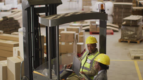 african american male and female workers wearing safety suits and using laptop in warehouse