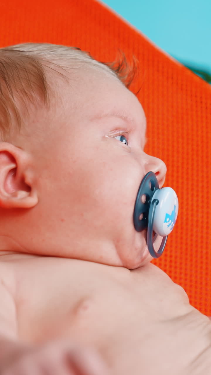 Cute blonde Caucasian infant with pacifier. Baby boy resting in the orange chair. Close up.. Vertical video