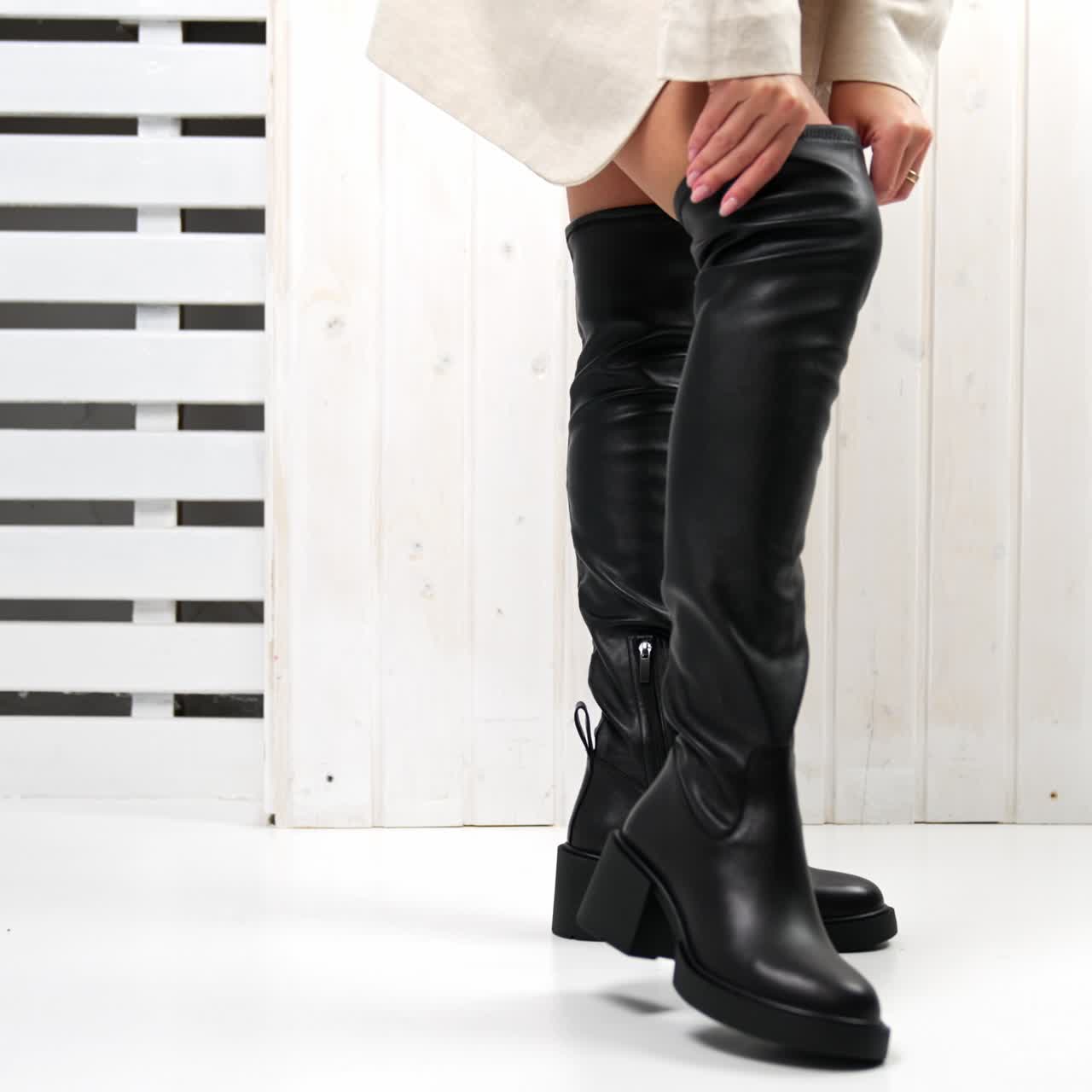 Girl in white mini skirt walking in high black boots. Model pulls the top of the footwear and demonstrates boots to the camera. White backdrop