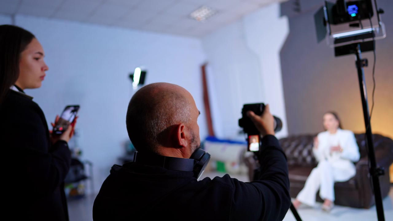 Camera Operator Filming Interview In Photo Studio. Camera operator films seated woman on couch while assistant monitors setup under studio lights.