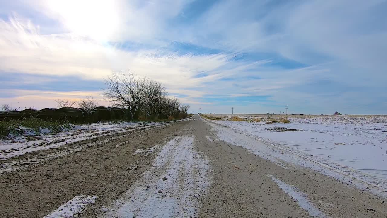 2X, POV through the rear window while driving on a snow covered gravel road past empty fields on a sunny winter afternoon; there is a light dusting of snow in the ditches; double time, point of view