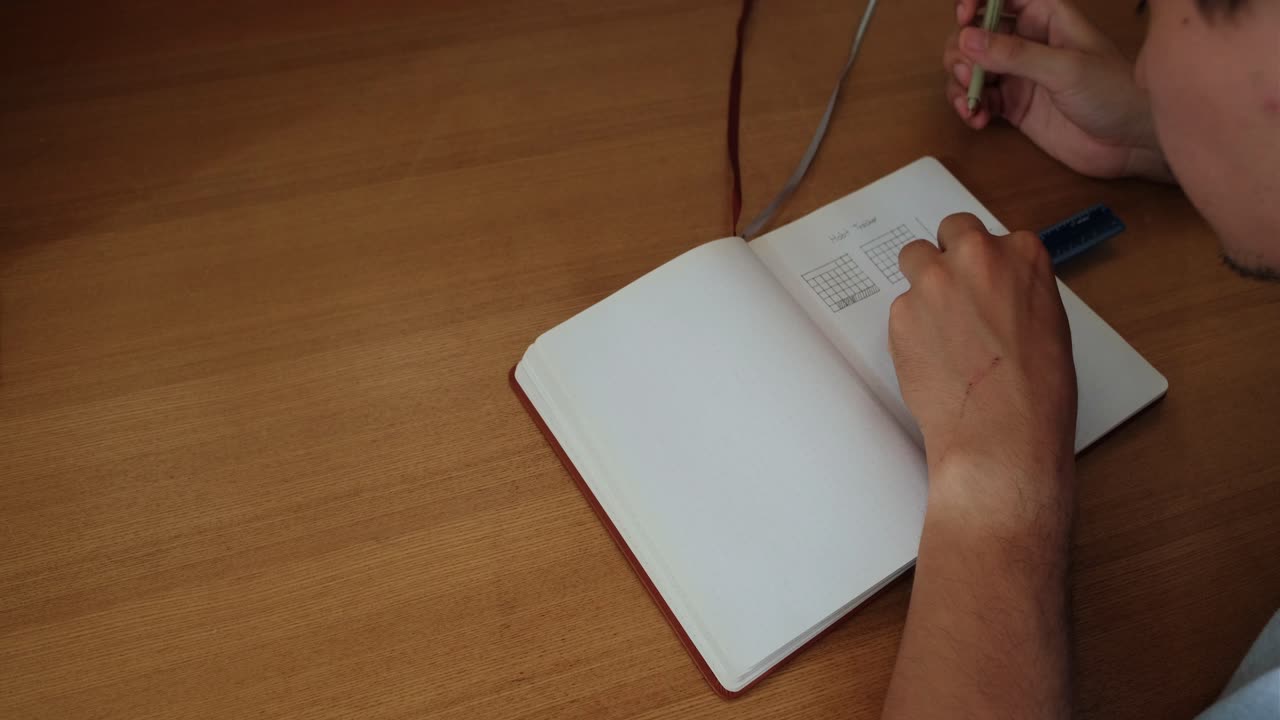 Medium overhead shot of a young man writing up lines for a habit tracker journal atop wooden desk