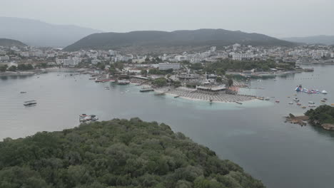 Drone-shot-flying-over-the-water-to-the-small-islands-in-front-of-Ksamil-Albania-in-the-morning-on-a-cloudy-grey-day-with-boats-around-and-haze-in-the-sky-LOG