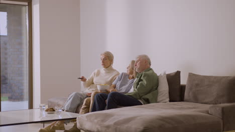 group of three senior people watching tv and talking to each other while sitting on sofa at home