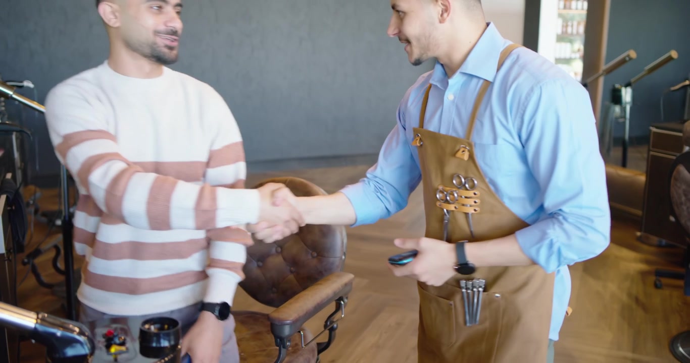 Cliente uomo, barbiere con grembiule che tiene il telefono, tocca il lettore, si stringono la mano e si prepara per il prossimo cliente.