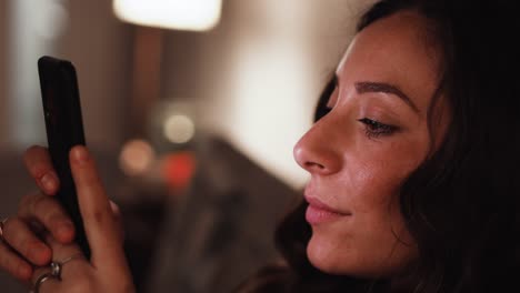 Young-Italian-woman-using-a-smart-phone-scrolling-and-smiling-while-sat-on-the-sofa-at-home