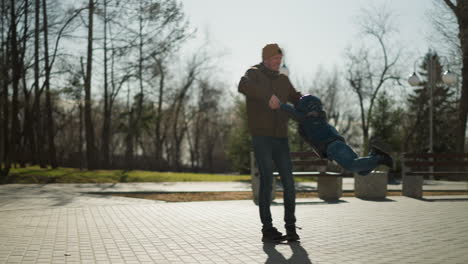 a close up of a father and son playing outdoor as the father spin his son playfully, with trees and benches in the background