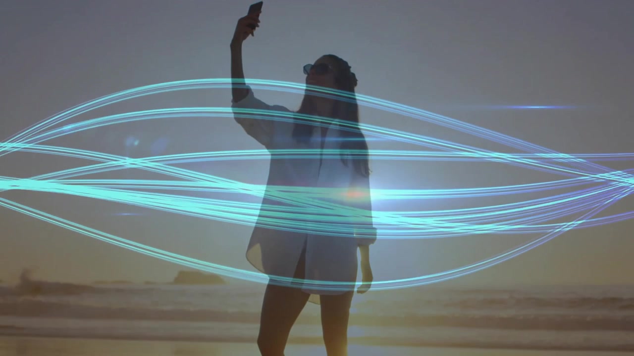 Mulher levantando o telefone acima da cabeça, enquadrando uma foto do pôr do sol com fitas de luz turquesa tecnológica varrendo a praia