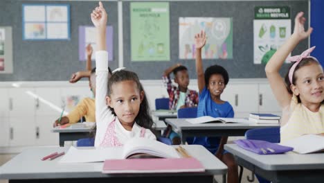 Fröhliche,-Vielfältige-Schulkinder-An-Schreibtischen,-Die-Im-Klassenzimmer-Der-Grundschule-Die-Hände-Heben