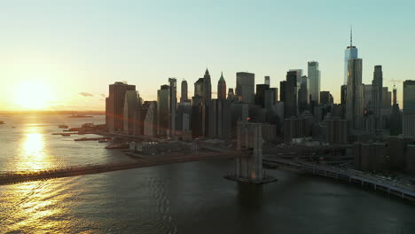 Forwards-fly-above-river.-Aerial-view-of-Brooklyn-bridge-against-bright-sunset-sky.-Skyscrapers-in-Financial-District.-Manhattan,-New-York-City,-USA