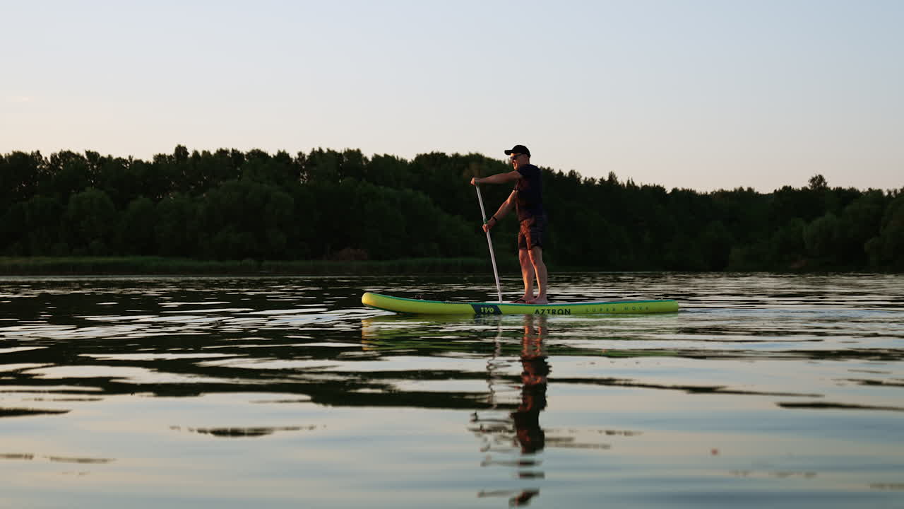 Man wearing black cap, t-shirt and shorts practices sup boarding. Nice active tranquil time spending on the river in summer evening.