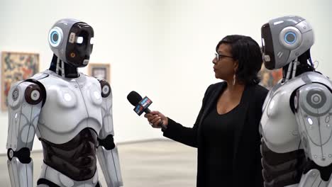 a groundbreaking interview: a woman engages with two advanced robots in a contemporary art space, exploring the intersection of technology and humanity