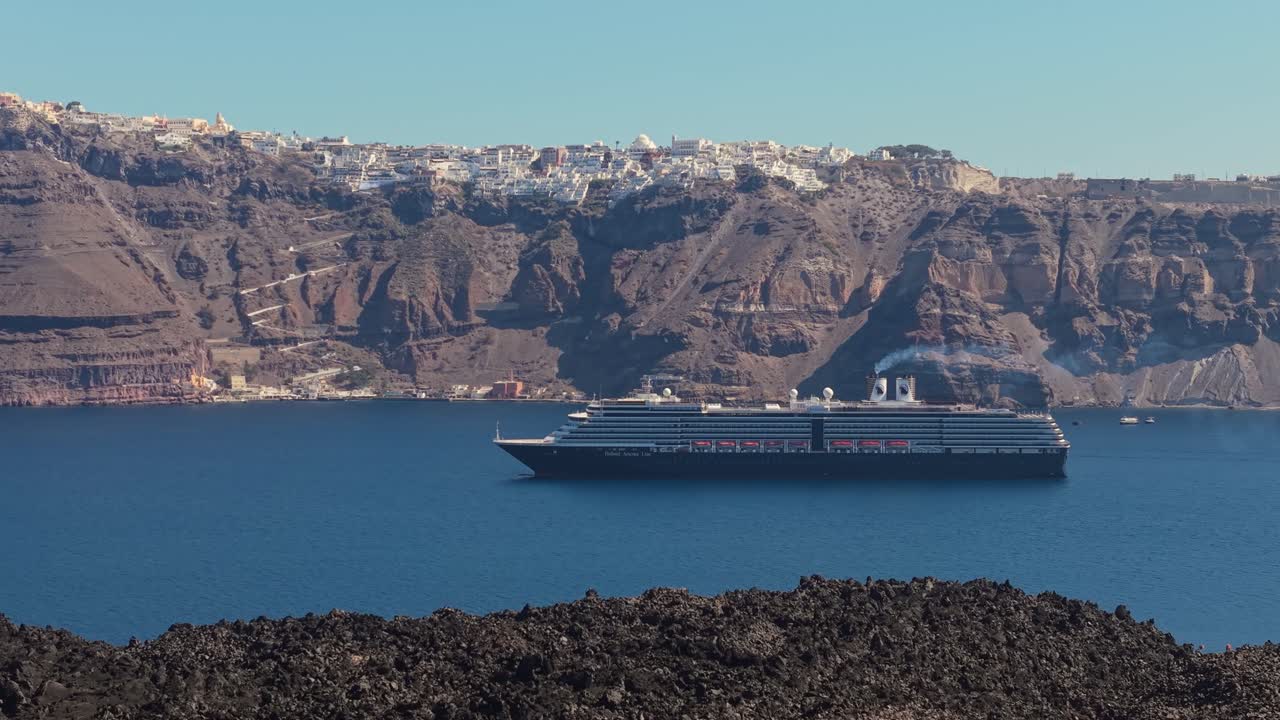 Sidelæns droneoptagelse af et krydstogtskib i Santorinis caldera med vulkansk lavajord i forgrunden og dramatisk ægæisk havudsigt i baggrunden.