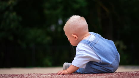 blond cute kid sitting on the ground outdoors in summer. little blond baby plays with sand. blurred backdrop.