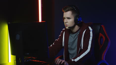 close up view of young serious man with headphones and playing a game on the computer in a room with colorful neon lamps on the wall 6