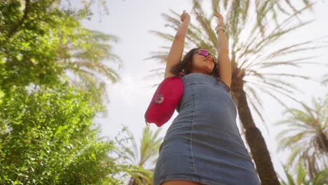 young woman in denim dress and sunglasses posing under palm trees