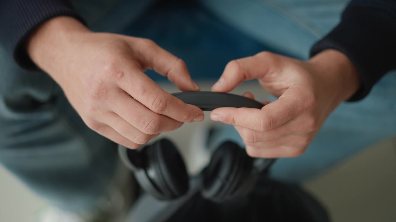 Närbild på händer som håller och justerar svarta hörlurar, med fokus på texturdetaljer. Händerna manipulerar hörlursbandet med synliga hudtexturer som förhårdnader eller ärr. Bakgrunden visar...