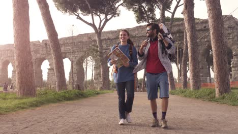 young couple backpackers tourists walking with map guide near roman aqueduct arches in parco degli acquedotti park ruins in rome at sunrise with guitar and sleeping bag slow motion steadycam