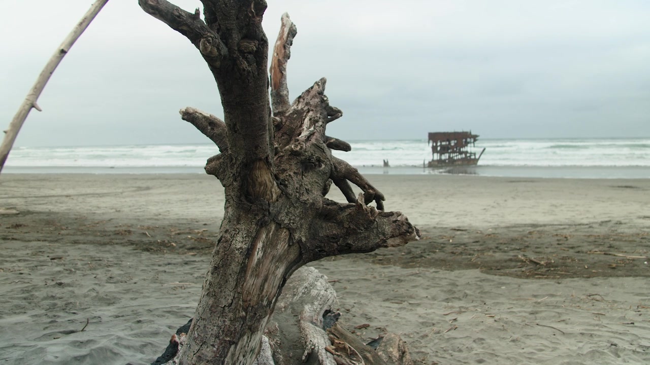 Uma tomada ampla em câmera lenta de um grande pedaço de madeira flutuante em primeiro plano com o naufrágio de Peter Iredale fora de foco ao fundo em um dia nublado de verão na praia costeira do Oregon