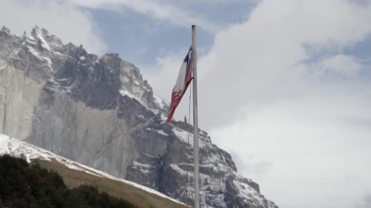 Působivý, symbolický snímek chilské státní vlajky (La Estrella Solitaria) vlající na pozadí členitých, zasněžených horských štítů národního parku Torres del Paine v Patagonii.