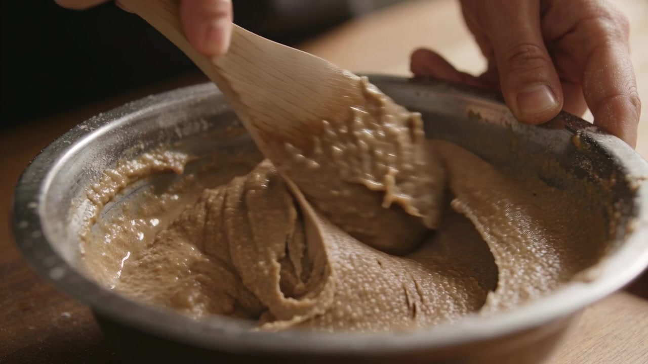 Stirring hands dark cuff steadying metal bowl at kitchen counter scraping spatula to smooth batter