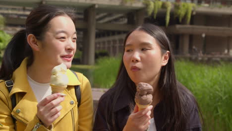 two smiling young female friends meeting and eating ice cream outdoors in park together