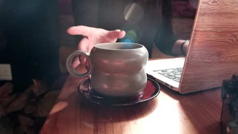 Woman-sitting-in-a-coffee-shop-working-on-her-laptop-and-drinking-a-hot-beverage-with-beautiful-soft-sun-flares