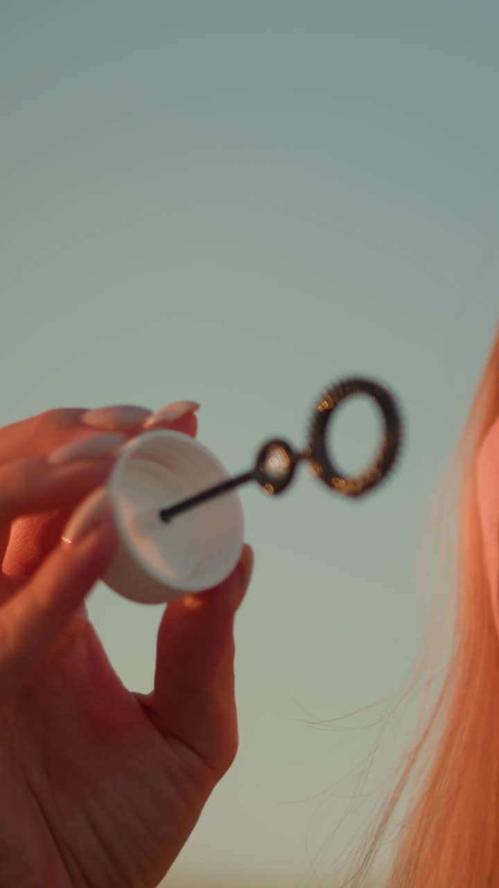 머리에 선글라스를 착용한 백인 여성이 해가 지는 동안 야외에서 거품을 불어넣고, 빛이 그녀의 얼굴에 따뜻하게 반사되어 거품 지이를 들고있는 동안 그녀의 특징을 조명합니다.