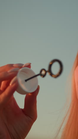 primer plano de una mujer blanca con gafas de sol en la cabeza soplando burbujas al aire libre durante la puesta del sol, la luz del sol se refleja cálidamente en su cara, iluminando sus rasgos mientras sostiene la varita de burbujas