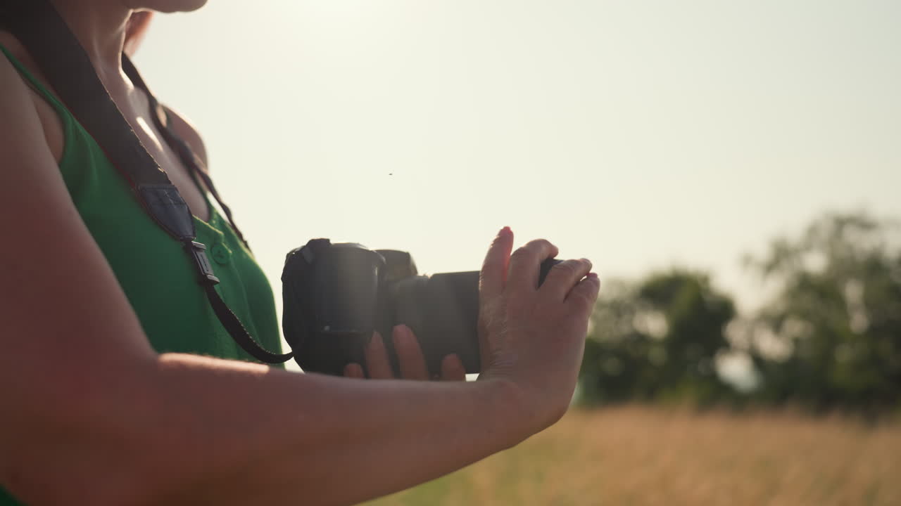 Tourist with closed eyes standing in golden field wearing camera strap around neck after removing lens cover from camera under soft sunlight during peaceful outdoor moment in summer countryside