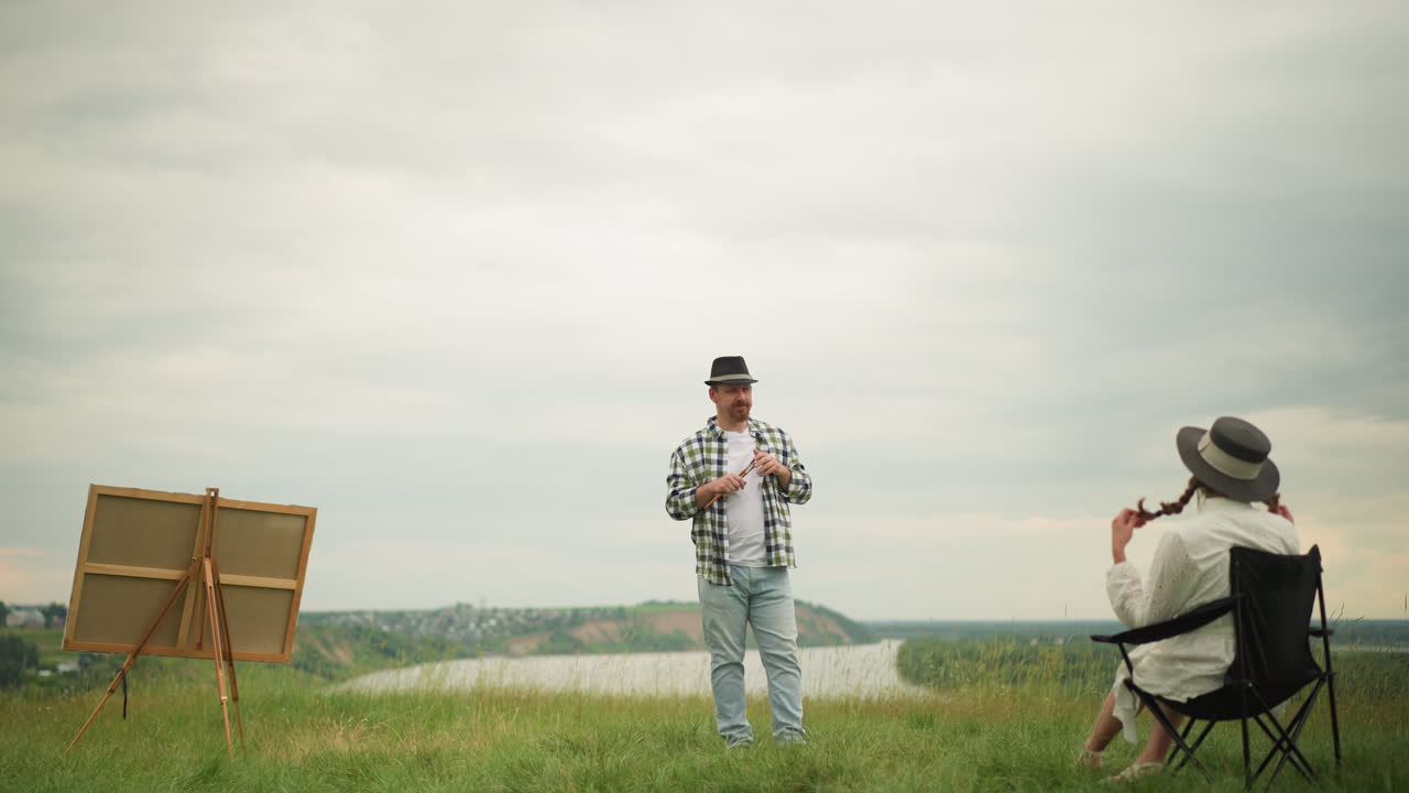 A handicrafter, holding a brush and wearing a checked shirt, observes a woman in a white dress and hat sitting on a chair in a grassy field by a serene lake