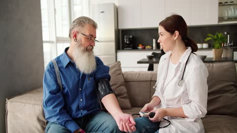 A-brunette-woman-in-a-white-coat-a-doctor-expresses-her-opinion-about-the-pulse-after-examining-an-elderly-man-with-gray-hair-and-a-lush-beard-in-glasses-in-a-blue-shirt-at-home-on-the-sofa-and-they-are-happy