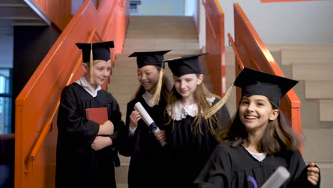 Feliz-Estudiante-De-Preescolar-Con-Toga-Y-Birrete-Sosteniendo-Un-Libro-Y-Mirando-La-Cámara-Mientras-Sus-Compañeros-De-Clase-Bajan-Las-Escaleras-En-La-Ceremonia-De-Graduación