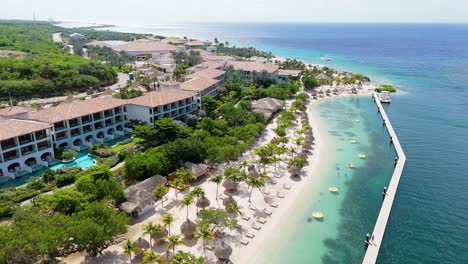 Panoramic-aerial-overview-of-lounge-chairs-below-palm-trees-and-tropical-umbrellas-at-beach-side-resort-Curacao