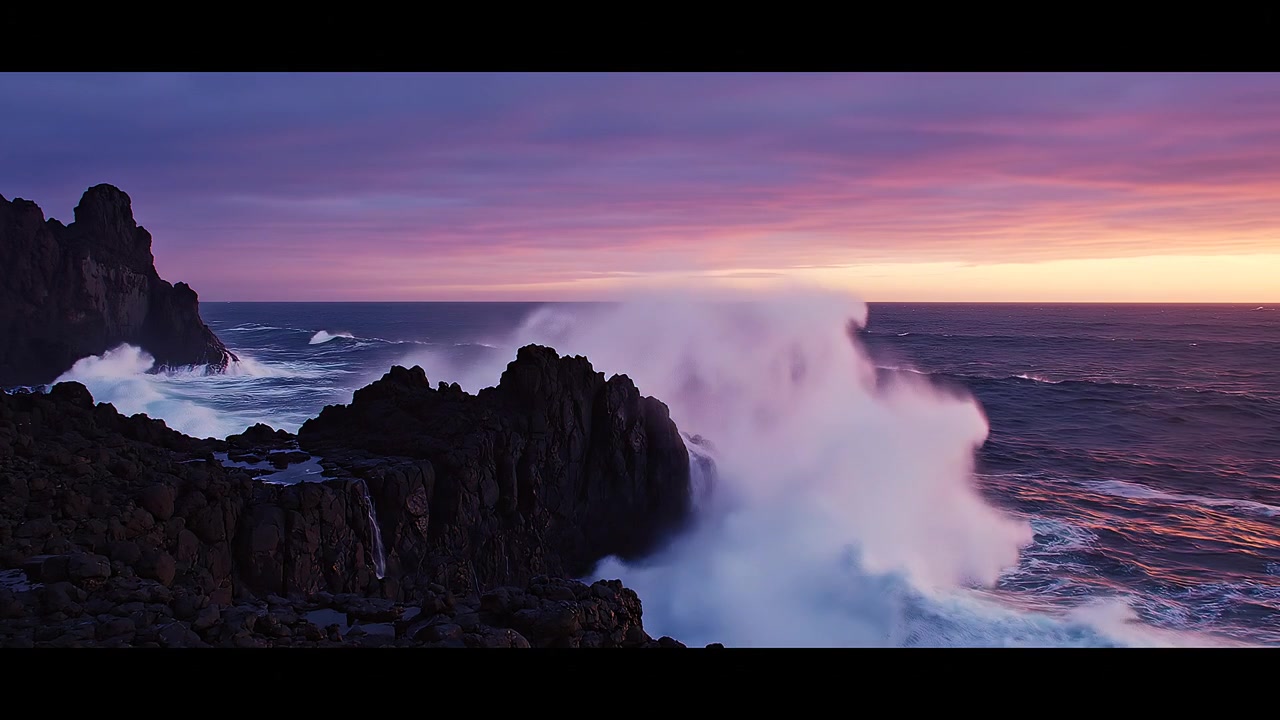Onde dell'oceano che si infrangono contro scure rocce costiere al tramonto viola