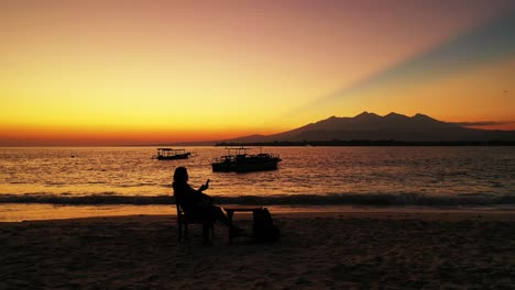 Young-man-relaxing-as-a-beautiful-orange-and-yellow-sun-sets-behind-distant-mountains-and-reflects-into-the-ocean