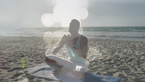 animation of glowing lights over african american man doing yoga meditation on beach