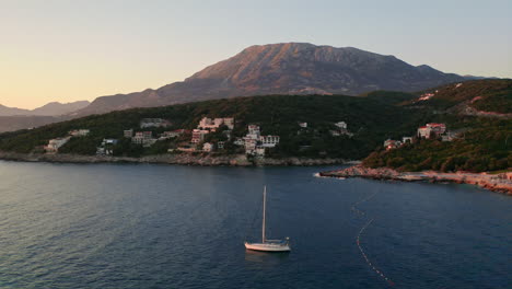 static aerial shot of a yacht anchoring in a bay in montenegro in the evening with a small coastal city and a large mountain in the background and a clear sky above